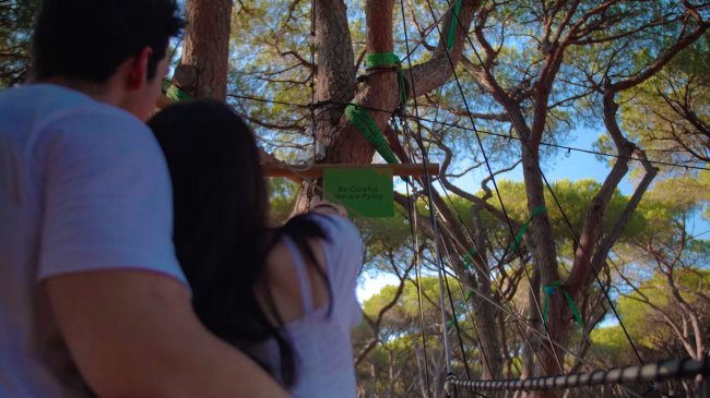 Due persone osservano un percorso avventura tra gli alberi, con una targhetta verde appesa.