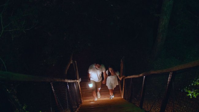 Una coppia cammina su una passerella di legno nella foresta di notte, con una lanterna.