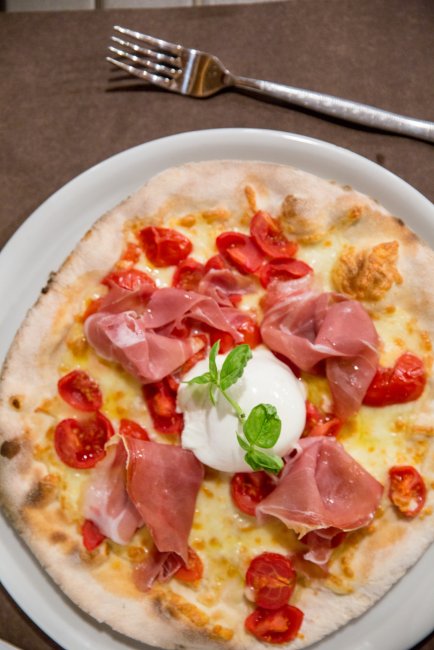 Pizza con prosciutto, pomodorini e burrata su un piatto bianco, accompagnata da una forchetta.