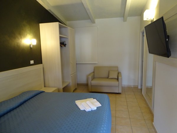Camera con letto blu e un divano beige. TV a parete e guardaroba aperto.
