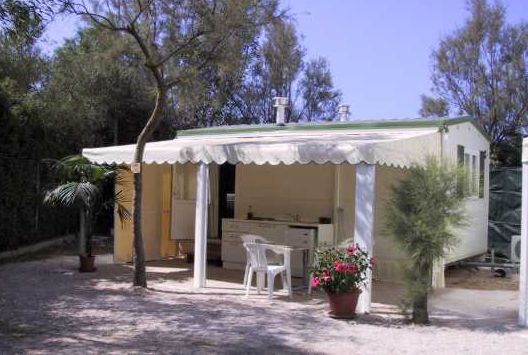 Una casa mobile con terrazza e sedie, immersa tra alberi e ghiaia.