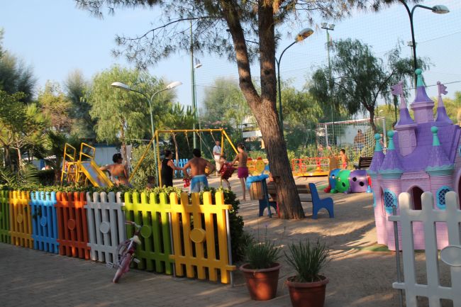 Area giochi colorata per bambini con staccionata, scivolo e altalene tra gli alberi.