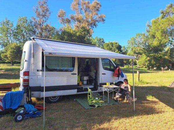Furgone bianco con tenda parasole, tavolo e sedie da campeggio all'aperto in un prato.