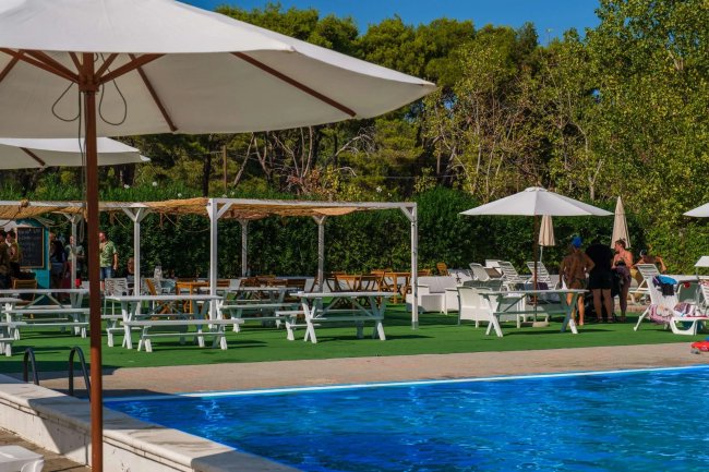 Una piscina con ombrelloni bianchi e tavoli da picnic su un prato verde circondato da alberi.