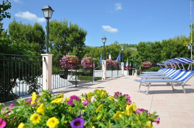 Una terrazza con sdraio blu e bianche, fiori colorati e lampioni, circondata da alberi verdi.