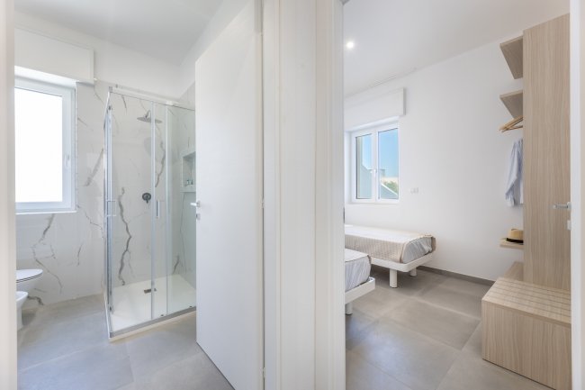 Bagno e camera con letti singoli, vista da un corridoio.