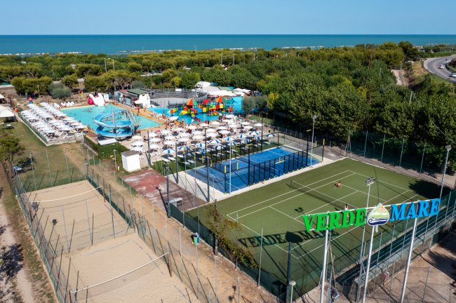 Vista aerea di un centro sportivo con piscine, campi sportivi e ombrelloni vicino al mare.