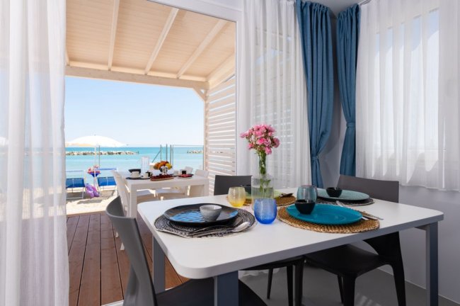 Sala da pranzo con vista mare, tavolo apparecchiato, fiori rosa, tenda blu.