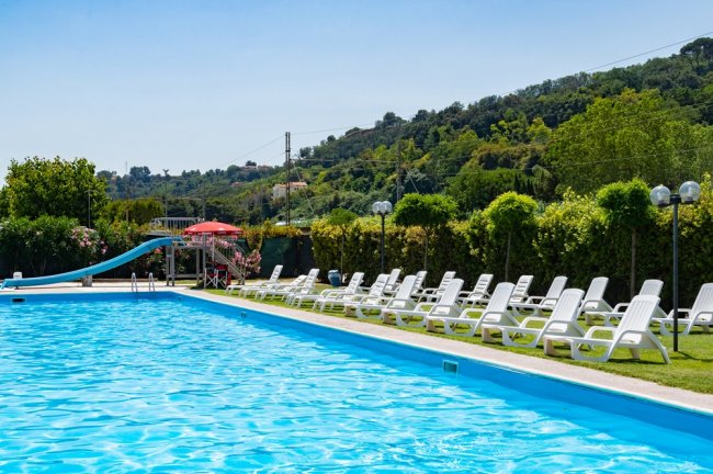 Piscina con scivolo blu, sdraio bianche su prato, colline sullo sfondo.