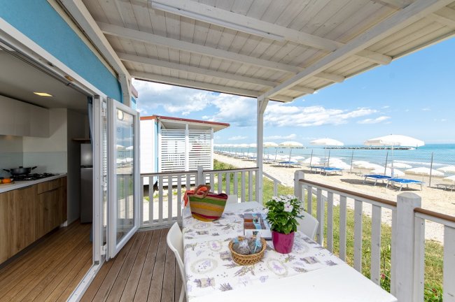 Terrazza con vista sul mare, tavolo apparecchiato e fiori, spiaggia con ombrelloni bianchi.