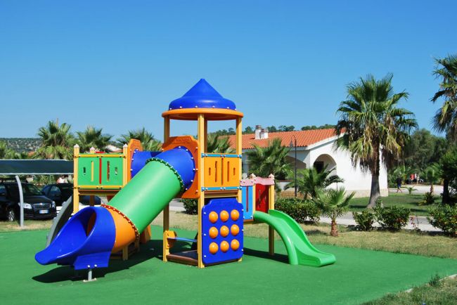 Area giochi colorata con scivoli e torrette su erba sintetica, cielo limpido e palme sullo sfondo.