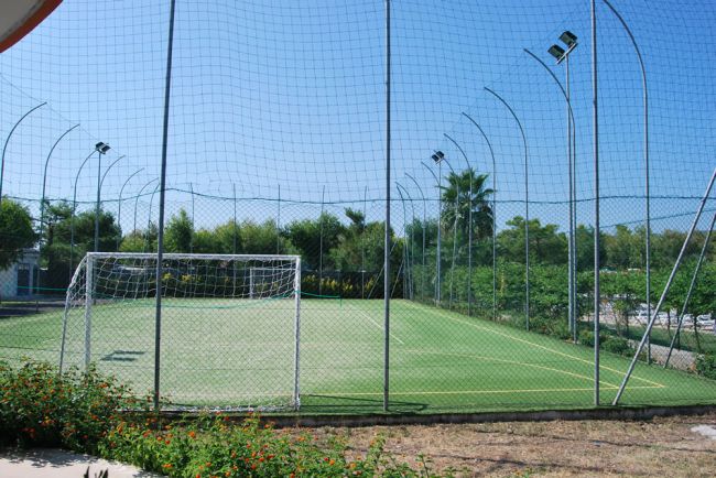 Campo da calcetto con rete intorno, illuminato da fari, circondato da alberi.