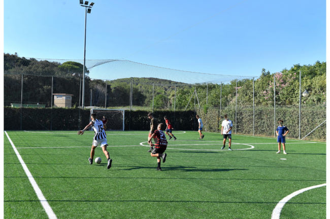 Calciatori in campo sintetico recintato, cielo sereno.