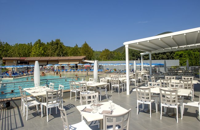 Ristorante all'aperto con vista sulla piscina affollata e montagne sullo sfondo.
