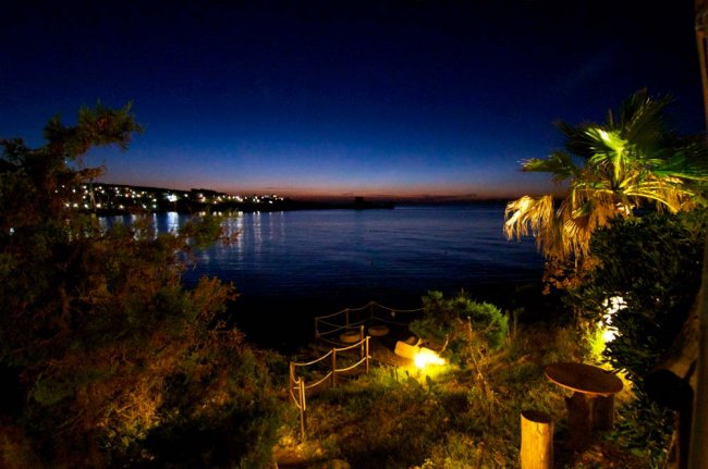 Vista notturna di una baia illuminata con cielo scuro e vegetazione in primo piano.