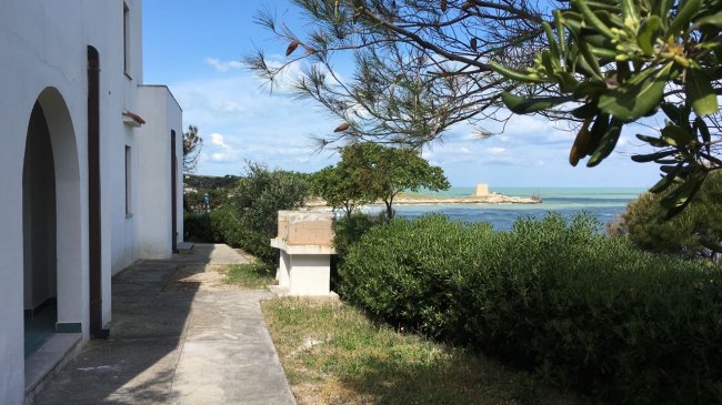Vista su un vialetto costiero con edificio bianco, cespugli e mare azzurro in sottofondo.