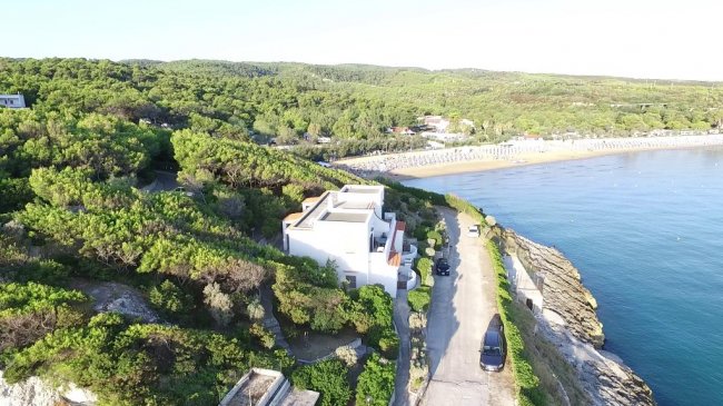 Veduta aerea di un edificio bianco sulla costa circondato da vegetazione e mare.