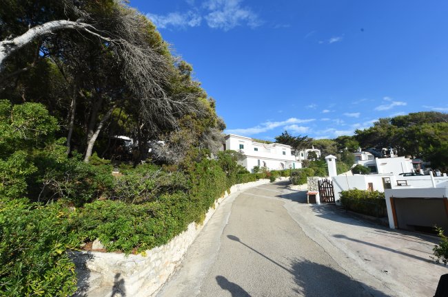 Strada in salita con case e vegetazione ai lati sotto un cielo blu.