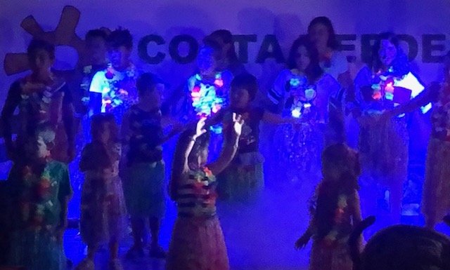 Bambini su un palco sotto luci colorate, indossano abiti festivi.