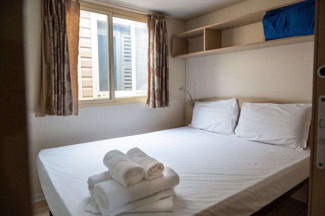 Camera da letto semplice con letto matrimoniale, lenzuola bianche e finestre con tende.