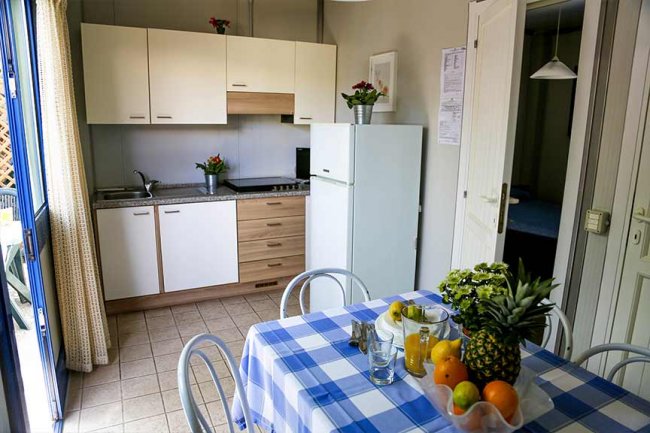 Cucina con tavolo apparecchiato e frutta, mobiletti bianchi e frigorifero.