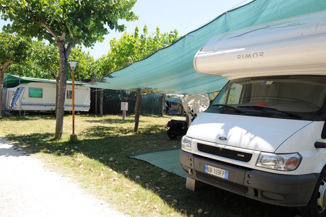 Campeggio con camper bianco parcheggiato sotto una tettoia verde e roulotte sullo sfondo.