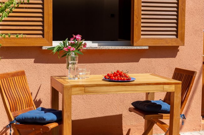 Tavolo di legno con piatto di pomodori, due sedie e vaso di fiori, davanti a una finestra.