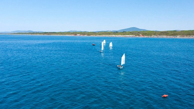 Barche a vela sul mare blu, con costa verdeggiante e montagne sullo sfondo.
