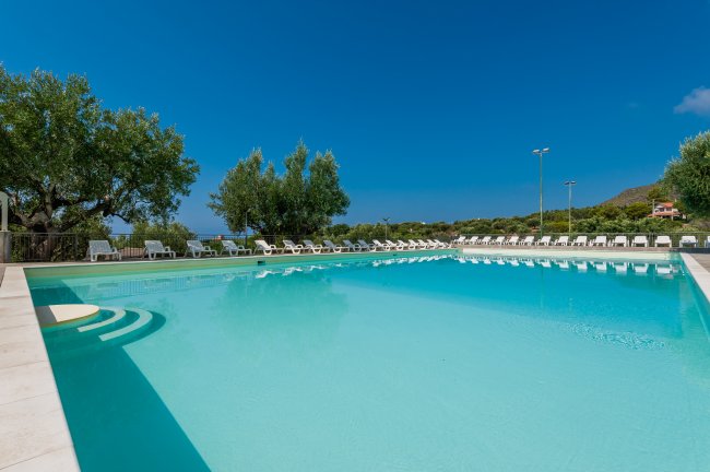 Piscina con gradini, circondata da lettini e ulivi, sotto un cielo sereno.