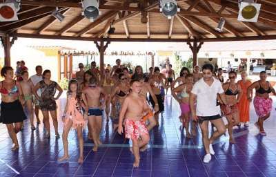 Gruppo di persone che ballano in costume su una pista blu sotto un tetto di legno.