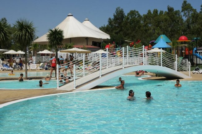 Piscina con un piccolo ponte bianco, bagnanti e ombrelloni, con un grande gazebo sullo sfondo.