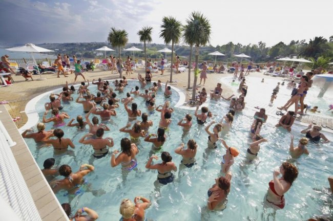 Persone in piscina affollata con palme, sotto un cielo nuvoloso.