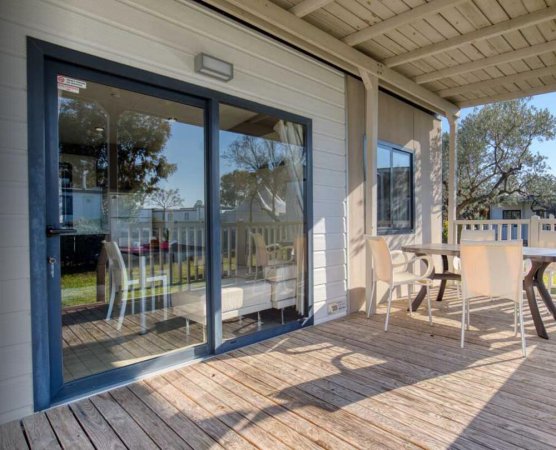 Terrazza con tavolo e sedie su pavimento in legno, vista giardino e porta finestra riflettente.