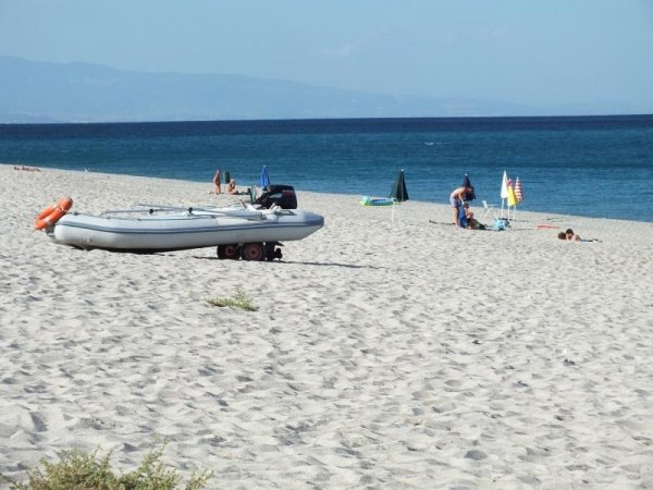 Gommone su spiaggia sabbiosa, mare e bagnanti sullo sfondo.