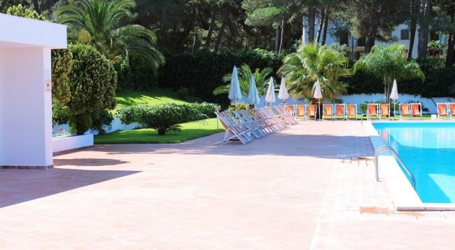 Area piscina con sdraio e ombrelloni bianchi, circondata da alberi.