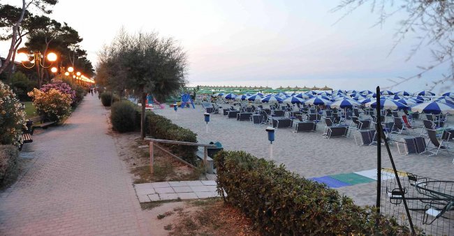 Sentiero accanto a una spiaggia con ombrelloni blu e bianchi all'alba; luci lungo il percorso.