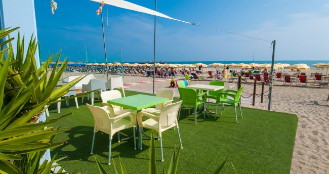 Tavoli e sedie verdi su prato sintetico vicino a spiaggia sabbiosa.