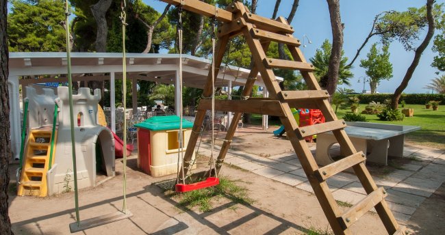 Area giochi con altalene, scivolo in plastica e tavolo da ping-pong tra gli alberi.
