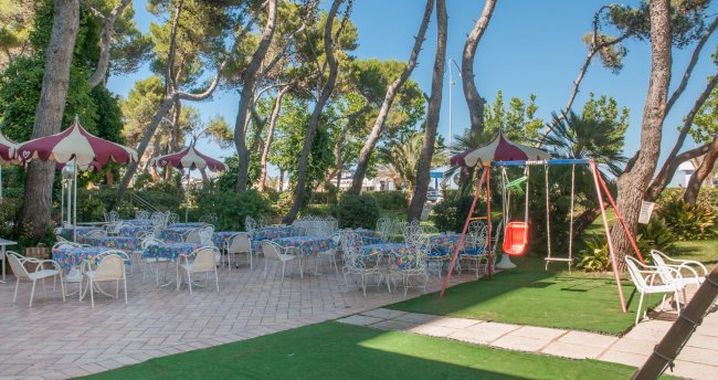 Giardino con tavoli e sedie, altalena rossa e alberi alti.