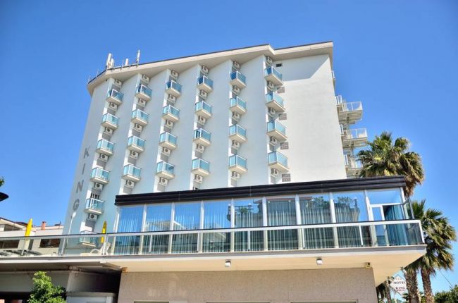 Edificio moderno bianco con balconi, circondato da palme, cielo blu sullo sfondo.
