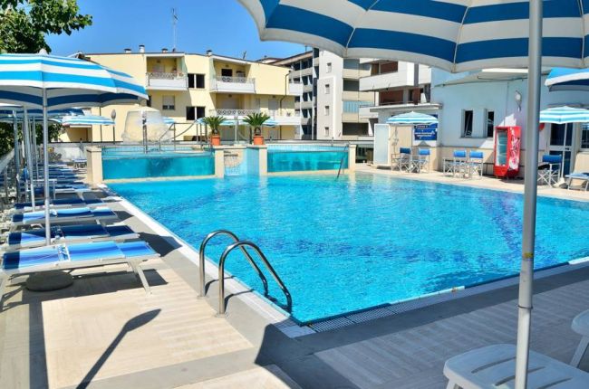 Piscina azzurra con lettini e ombrelloni a strisce blu e bianche, edifici sullo sfondo.