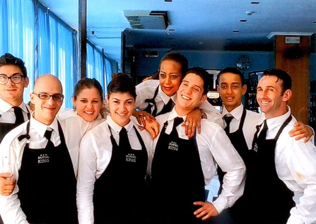 Gruppo di camerieri sorridenti in uniforme con grembiule nero, posano insieme.