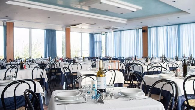 Una sala da pranzo con tavoli apparecchiati e sedie nere, tende blu alle finestre.