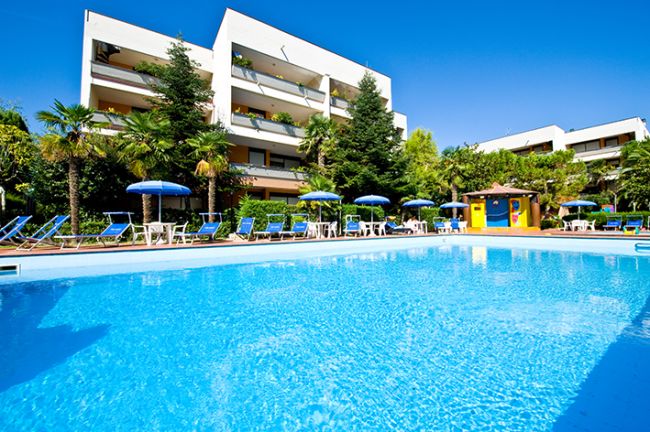 Piscina circondata da lettini blu e ombrelloni, edificio bianco a più piani sullo sfondo.