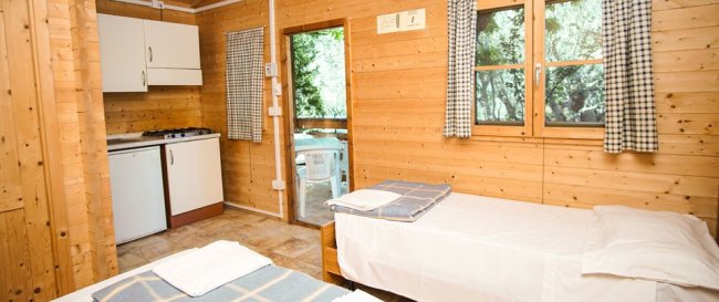 Interno di una cabina in legno con due letti, angolo cottura e vista su una porta finestra.