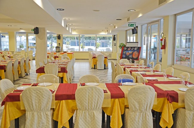 Sala da pranzo con tavoli apparecchiati, tovaglie rosse e gialle.