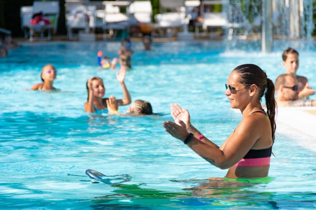 Un'istruttrice in piscina guida una lezione di aquagym; le persone seguono i movimenti.