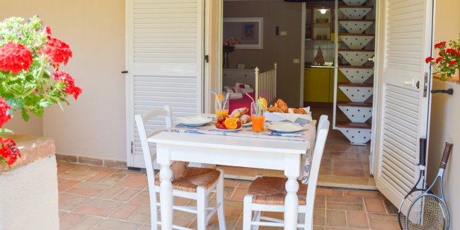 Tavolo da pranzo all'aperto con sedie bianche, colazione servita e fiori rossi.