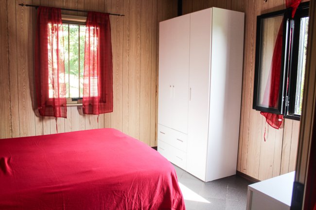 Camera da letto con letto matrimoniale rosso, armadio bianco e tende rosse. Pareti in legno chiaro.
