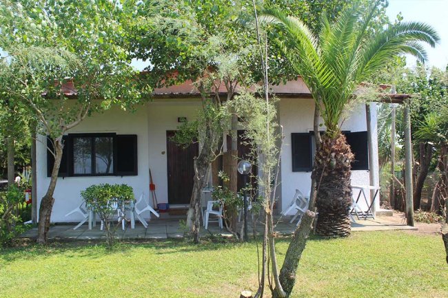 Casetta bianca con giardino, circondata da alberi e con sedie bianche sul patio.